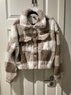 Rutig teddyjacka i beige och vitt - Supermysig teddyjacka med stora rutor i beige och vitt. Jackan har krage, knäppning framtill och två bröstfickor. 