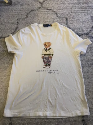 Vit Polo Bear t-shirt från Ralph Lauren - Vit t-shirt från Polo Ralph Lauren med ikoniskt Polo Bear-tryck på bröstet. Modellen är custom slim fit och har rund hals och korta ärmar. Tillverkad i mjuk bomull för en skön känsla. Perfekt statement-plagg för dig som gillar klassisk stil med en twist. 