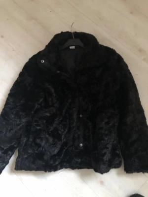 Svart pälsjacka med krage - Säljer en svart pälsjacka med mjuk och fluffig känsla. Jackan har en klassisk krage och stängs med knappar framtill. Perfekt för dig som vill ha en cool och varm look under kyliga dagar.