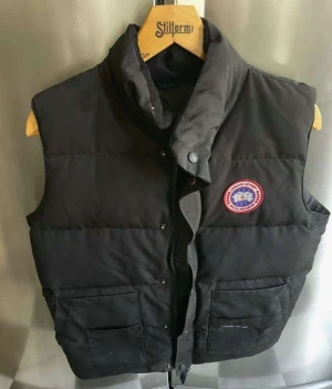 Svart dunväst från Canada Goose M - Svart dunväst från Canada Goose med klassisk logga på bröstet. Västen har hög krage, två stora fickor framtill och stängs med både dragkedja och tryckknappar. Perfekt för lager-på-lager under kyliga dagar. Fodrad med dun för extra värme. Äkta vara.