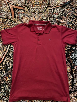 Vinröd piké från Tommy Hilfiger - Snygg vinröd pikétröja från Tommy Hilfiger med klassisk krage och två knappar framtill. Pikén har ett litet broderat logomärke på bröstet och diskreta svarta detaljer på kragen. Tillverkad i mjuk bomull som känns skön mot huden. Perfekt för en chill och stilren look. Ingen fri frakt 