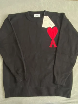 Svart stickad tröja från Ami med hjärta - Svart stickad tröja från Ami Alexandre Mattiussi med rund hals och ribbade muddar. På bröstet finns ett stort rött hjärta med bokstaven A. Tröjan har lång ärm och är perfekt för dig som gillar stilrena detaljer med en twist.