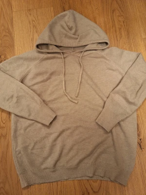 Beige stickad hoodie med huva - Mysig beige hoodie med stickad känsla och huva. Tröjan har långa ärmar, snörning vid halsen och ribbade muddar vid ärmslut och nederkant. Perfekt för chill dagar och när du vill ha det extra bekvämt.