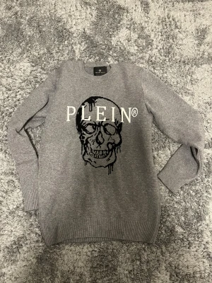 Grå stickad tröja med döskalle från Philipp Plein - Säljer en grå stickad tröja från Philipp Plein i storlek L. Tröjan har ett stort döskallemotiv i svart och vitt framtill samt tydlig logotyp. Rund hals och ribbade muddar vid ärmslut och nederkant. Perfekt för dig som gillar edgy streetstyle.