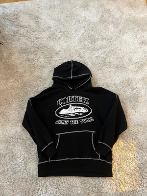 Svart Corteiz hoodie med vit print - Svart hoodie från Corteiz med vit kontrastsöm och stor vit print på bröstet med texten 'Corteiz Rules The World' och motiv. Tröjan har huva, magficka och långa ärmar. Materialet är mjuk bomull, perfekt för chill dagar.