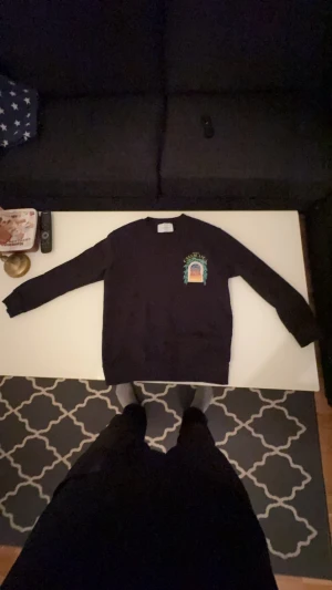 Svart sweatshirt från Casablanca - Svart sweatshirt från Casablanca med färgglad tryckt grafik på bröstet och ryggen. Motivet visar en byggnad med gröna blad och texten 'Avenida Casablanca' i gult. Tröjan har rund hals och långa ärmar, perfekt för dig som gillar unika prints och streetwear. Säljer denna underbara tröja från Cassablanca vill sälja den då jag har råkat krympa till den originals storleken är storlek S men funkar nu som storlek XS. Säljer den för 1000 kr men pris kan diskuteras vid snabba affärer! 