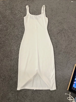 Vit långklänning med slits - Snygg vit långklänning med breda axelband och figurnära passform. Klänningen har en enkel, stilren design och en slits baktill för extra rörelsefrihet. Den har små flecks på fram och baksida 