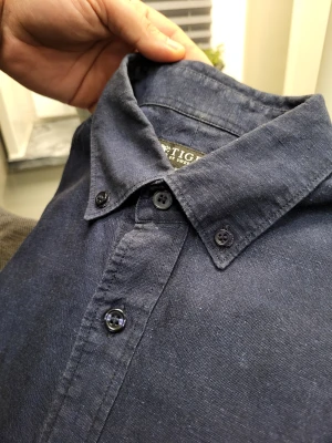 Mörkblå skjorta från Tiger of Sweden M - Snygg mörkblå skjorta från Tiger of Sweden i slim fit. Skjortan har klassisk krage med knappar, långa ärmar och knäppning framtill välanvänd kolla på sista bilden.Tillverkad i ett mjukt linnetyg som ger en avslappnad men stilren vibe. Perfekt för dig som gillar enkel och clean stil.