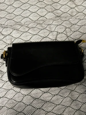 Svart axelväska i skinn med gulddetaljer - Snygg svart axelväska i slätt skinn med stilrena linjer och guldfärgade metalldetaljer. Väskan har ett lock med mjukt böjd kant och praktisk axelrem. Perfekt för dig som gillar minimalistisk och klassisk stil.