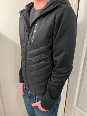 Svart quiltad jacka Sail Racing - Riktigt sjyst svart jacka från Sail Racing med huva och dragkedja. Ärmarna är släta och i mjukare material, medan kroppen är i ett glansigare. 