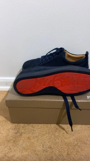 Marinblå sneakers från Christian Louboutin - Snygga Marinblå sneakers från Christian Louboutin med klassisk röd sula och diskret logga undertill. Skorna har rund tå, snörning och är tillverkade i mocka med beige innersida. Perfekta för dig som vill ha en lyxig och stilren look.