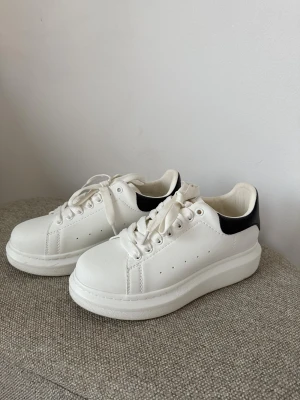 Alexander McQueen vita sneakers - Säljer ett par ikoniska vita sneakers från Alexander McQueen med svart hälparti och guldfärgad logga. Skorna har tjock platt sula, rund tå och klassisk snörning. Tillverkade i skinn med stilren design som ger en clean och modern vibe.
