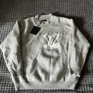 Grå stickad tröja Louis Vuitton - Supersnygg grå stickad tröja från Louis Vuitton med stort vitt LV-logo framtill. Tröjan har rund hals, ribbade muddar och lång ärm. Perfekt för dig som gillar exklusiva märken och vill ha en clean look.