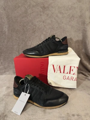 Valentino rockrunners  - Valentino Garavani Rockrunner Sneakers – Svart / Strl42 men passar 42,5 Säljer ett par äkta Valentino Garavani Rockrunner-sneakers i svart. Skorna är i mycket fint skick och kommer komplett med originalkartong, dustbag, taggar och originalfyllning – perfekt både för användning och som samlarobjekt.  Skick: ✔ Mycket bra skick  Detaljer: 	•	Modell: Valentino Garavani Rockrunner 	•	Färg: Svart med mörkbruna detaljer 	•	Materialmix med läder, mocka och mesh 	•	Klassiska pyramidnitar vid hälen 