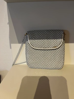 Vit och svart axelväska med kedja - Snygg axelväska i vitt med svart rutmönster och gulddetaljer. Väskan har en smidig kedjerem och magnetlås. Perfekt storlek för mobilen och det viktigaste. Stilren och modern design som sticker ut.