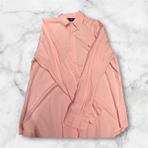 Rosa skjorta från Ralph Lauren - Snygg rosa skjorta från Ralph Lauren med klassisk krage och knappar framtill. Skjortan har lång ärm och en liten broderad logga på bröstet. Perfekt för dig som gillar stilrena och färgglada plagg.