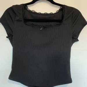 Svart ribbad topp med spets och rosett - Snygg svart ribbad topp med korta ärmar och fyrkantig urringning. Dekorerad med spetskant och en liten rosett framtill. Perfekt för dig som gillar en söt men stilren look. Jättebra skick, inga synliga slitningar. Knappt använd.