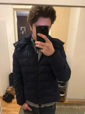 Superdry dunjacka   - Säljer en mörkblå dunjacka från Superdry med avtagbar huva och dragkedjor på fickorna. Jackan har ett broderat Superdry-märke på ärmen och är vadderad för extra värme. Perfekt för kalla dagar.