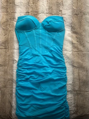 Turkos axelbandslös långklänning - Snygg turkos axelbandslös långklänning med vadderade kupor och markerade sömmar upptill. Klänningen har rynkade detaljer längs sidorna och är figurnära med en tight passform. Perfekt för dig som vill sticka ut med en färgstark look.
