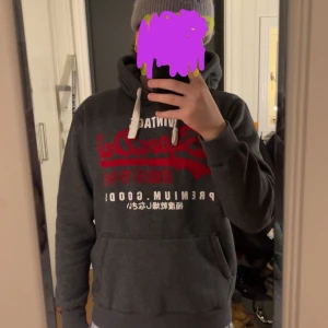 Mörkgrå Superdry hoodie med tryck - Säljer en mörkgrå hoodie från Superdry med rött och vitt tryck på bröstet. Tröjan har huva med vita snören och en stor magficka. Perfekt för dig som gillar streetstyle och vill ha något bekvämt och snyggt.