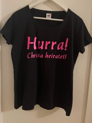 Svart t-shirt med rosa text Fruit of the Loom - Svart t-shirt från Fruit of the Loom med rund hals och korta ärmar. Framsidan har ett stort tryck i metallicrosa med texten 'Hurra! Christa heiratet!'. Modellen är Lady Fit och materialet är mjuk bomull. Perfekt för roliga tillfällen och statement.