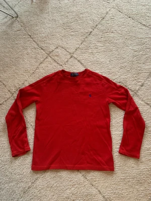 Röd långärmad tröja från Polo Ralph Lauren - Röd långärmad tröja från Polo Ralph Lauren med klassisk rund hals och blå broderad logga på bröstet. Tröjan har en lätt ribbad struktur och är perfekt för dig som gillar stilren och enkel design.