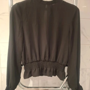 Svart blus med smock och volang - Svart blus med långa ärmar, smockdetaljer längst ner och vid ärmsluten samt volangkant. Tunnare tyg med rund halsringning och en loose fit. Perfekt för dig som gillar stilrena och trendiga plagg.