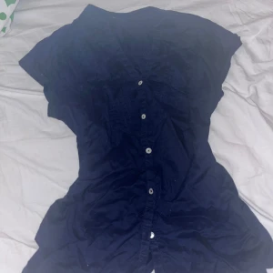 Mörkblå kortärmad skjorta Monoprix - Mörkblå skjorta från Monoprix Autre Ton i storlek 36. Skjortan har korta ärmar, v-ringning och knappar framtill. Snygga veckade detaljer på bröstet ger en cool touch. Perfekt för dig som gillar enkel men stilren look. Är mörkblå, ser mycket mörkare ut på sista bilderna pga dåligt ljus