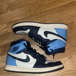 Nike Air Jordan 1 blå och vita sneakers( obsidian) - Säljer ett par Nike Air Jordan 1 sneakers i blått, vitt och svart. Klassisk high-top modell med svart swoosh, blå detaljer och vita paneler. Skorna har snörning och är tillverkade i skinn. Perfekta för dig som gillar streetwear och vill sticka ut. Säljs pågrund av att den inte passar längre, använd i nyskick. Pris går att diskuteras vid snabbaffär.