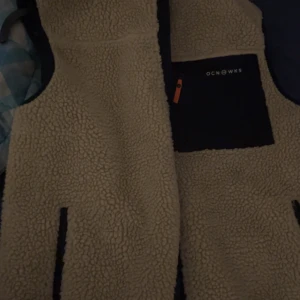 Beige fleeceväst från Ocean Works - Mysig beige fleeceväst från Ocean Works med kontrasterande svart bröstficka och dragkedja med orange detalj. Västen har två fickor framtill och är perfekt för lager-på-lager-stil under kyliga dagar.