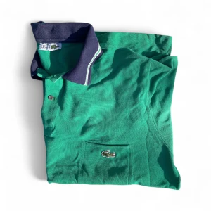 Grön lacoste piké - Snygg grön pikétröja från Lacoste med klassisk krokodillogga på bröstfickan. Tröjan har mörkblå krage med vita ränder och matchande detaljer på ärmsluten. Tillverkad i bomull, perfekt för chill dagar. Två knappar vid halsen för extra stil. Priset är absolut inte hugget i sten! Skriv om ni vill ha fler bilder eller har några frågor så ska jag försöka mitt bästa! ///Mvh Arvid