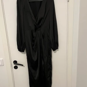 Svart omlottklänning från SHEIN - Snygg svart långklänning från SHEIN med omlottdesign och v-ringad hals. Klänningen har långa ärmar med lätt puff och är gjord i ett glansigt polyestermaterial som ger en elegant look. Perfekt för dig som gillar stilrena och trendiga plagg.