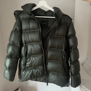 Mörkgrön pufferjacka från Massimo Dutti - Säljer en snygg mörkgrön pufferjacka från Massimo Dutti. Jackan har glansig yta, huva, två dragkedjefickor och kraftig dragkedja framtill. Perfekt för kalla dagar och ger en cool, modern look. Passformen är normal och jackan är vadderad för extra värme. Jag är ca 165 cm, och den sitter rätt så oversize på mig. 