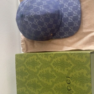 Blå keps från Gucci med GG-mönster - Snygg blå keps från Gucci med ikoniskt GG-mönster över hela ytan och en liten guldfärgad GG-detalj på sidan. Kepsen är tillverkad i ett slitstarkt bomullsmaterial och har klassisk form med böjd skärm. Perfekt för dig som vill ha en lyxig streetstyle-accessoar.