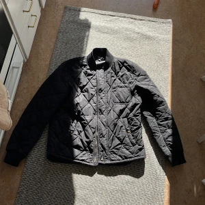 Svart quiltad bomberjacka - Quiltad svart bomberjacka med ribbad krage och muddar. Jackan har dragkedja och knappar framtill, samt fickor med tryckknappar. Snyggt rutmönster över hela jackan och långärmad modell. Perfekt för dig som gillar stilren och sportig look.