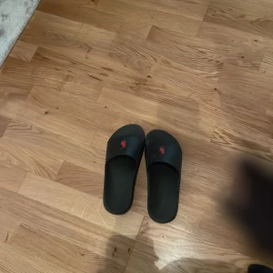 Svarta sandaler från Polo Ralph Lauren - Svarta sandaler från Polo Ralph Lauren med röd logga på ovansidan. De har en enkel och stilren design med platt sula och öppen tå. Tillverkade i syntetmaterial, perfekta för sommaren. Märket syns även på undersidan av sulan.