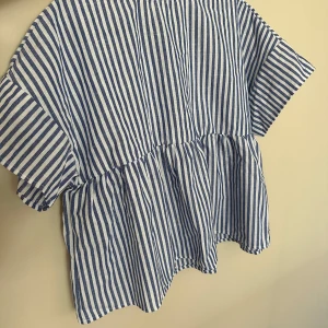 Randig blus från SHEIN i blåvitt - Superfin blus från SHEIN i blåvitt randigt mönster. Modellen är loose fit med korta ärmar och volang nedtill. Tillverkad i ett lätt och luftigt material som passar perfekt för varmare dagar. Stängs med knapp i nacken för en söt detalj.