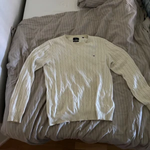 stickad GANT tröja - Vit kabelstickad tröja från GANT i premium bomull. Klassisk rund halsringning, ribbade muddar och diskret logga på bröstet. Perfekt för dig som gillar en clean och tidlös stil.