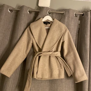 Beige Kappa från Vero Moda XS - Snygg beige Kappa från Vero Moda i storlek XS. Jackan har bred krage, knytskärp i midjan och raka ärmar. . Materialet är mjukt och känns lyxigt, passar perfekt till höst.