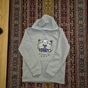 Grå Kenzo hoodie med tigertryck - Säljer en grå hoodie från Kenzo med ikoniskt tigertryck och Kenzo Paris-logga på bröstet. Tröjan har huva, långa ärmar och är tillverkad i mjuk bomull. Perfekt för dig som gillar streetstyle och vill sticka ut med ett coolt motiv.