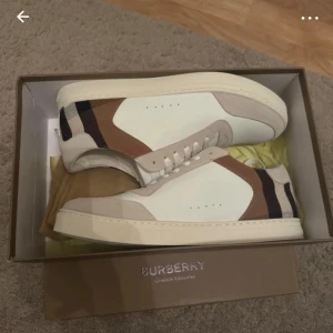 Burberry sneakers med klassiskt rutmönster - Snygga Burberry sneakers i vitt skinn med beige mockadetaljer och klassiskt rutmönster i brunt, svart och beige på hälen. Skorna har rund tå, platt sula och snörning framtill. Perfekta för dig som vill ha en stilren men ändå lyxig look.