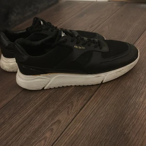 Svarta sneakers från Axel Arigato - Snygga svarta sneakers från Axel Arigato med vit chunky sula. Skorna har ovandel i skinn och mesh, med diskret guldfärgad logga på sidan. Klassisk snörning och sportig vibe, perfekta till streetwear eller casual outfits.
