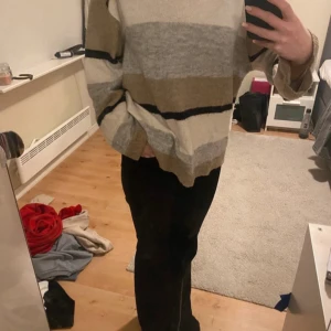 Randig oversized stickad tröja - Mysig oversized stickad tröja med breda ränder i beige, grått, vitt och svart. Tröjan har lång ärm och avslappnad passform, perfekt för lager-på-lager. Snygg att styla med jeans eller svarta byxor.