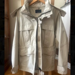 Beige trenchcoat från Slanta med huva - Snygg beige vindjacka från Slanta med stor huva, flera fickor och justerbara ärmslut. Jackan har dragkedja framtill och praktiska detaljer som gör den perfekt för blåsiga dagar. Passar dig som gillar funktionell och stilren design.