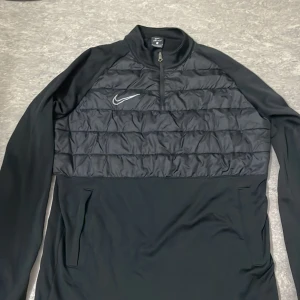 Nike dri fit - Snygg och funktionell svart långärmad tröja från Nike med Dri-FIT-teknologi. Tröjan har en dragkedja framtill och quiltad design på bröstet. Perfekt för träning eller vardagsbruk.