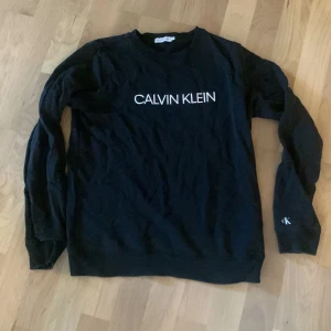 Svart Calvin Klein Jeans sweatshirt - Svart sweatshirt från Calvin Klein Jeans med vit logga framtill och liten 'ck'-detalj på ärmen. Klassisk rund halsringning och långa ärmar. Mjuk och skön i bomullsmix, perfekt för en clean och stilren look.