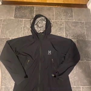 Svart vindjacka från Haglöfs  - Svart vindjacka från Haglöfs den är unisex men de står dam Xl men de för bådaJackan har huva, hel dragkedja framtill och två snedställda fickor med dragkedja. Materialet är lätt och smidigt, perfekt för blåsiga dagar. Klassisk Haglöfs-logga på bröstet.