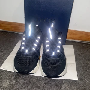 Svart vita dior b22 - Chunky svarta Dior sneakers med vit sula och reflekterande snören. Ovandelen är i mesh och skinn för en sportig och lyxig vibe. Snyggt loggad på sidan och tung design som sticker ut. Perfekta för dig som gillar statement-skor och streetstyle.