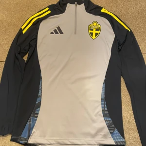 Svenska landslaget långärmad träningströja - Svenska landslagets långärmade träningströja från Adidas i slim fit. Grå front med svarta och gula detaljer, broderad landslagssköld och Adidas-logga. Tillverkad i funktionsmaterial (AEROREADY) som andas, perfekt för träning. Dragkedja vid halsen och klassiska ränder på axlarna.
