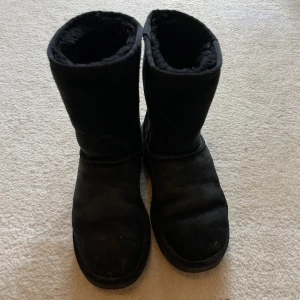 Svarta UGG boots i mocka - Mysiga svarta UGG boots i klassisk modell med rund tå och platt sula. Skorna är fodrade med får material på insidan och har en ovandel i mocka. Perfekta för kalla dagar när du vill hålla fötterna varma och bekväma. Väl använda men fortfarande fullt användbara och gör sitt jobb.